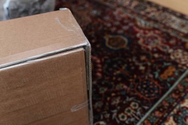 Tightly taped corner of a large cardboard box placed on a richly patterned carpet, highlighting durability and proper sealing for safe delivery. clipart