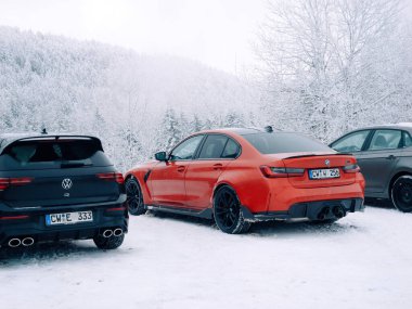 Black Forest, Germany - Jan 3, 2025: A red BMW M3 and black Volkswagen R parked in a snowy mountain lot, surrounded by frosted trees and a serene winter landscape. clipart