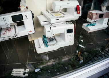 Aachern, Germany - Jan 3, 2025: A variety of sewing machines, including Brother, Bernette, and Pfaff models, displayed in a shop window with festive decorations and embroidery samples clipart