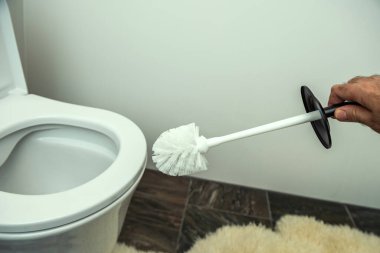 Siyah saplı beyaz bir tuvalet fırçası beyaz bir klozetin yanında, bej tüylü bir paspas ile döşenmiş..