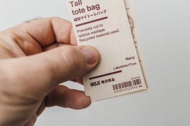 Paris, France - Oct 25, 2023: A close-up of a hand holding a MUJI tall tote bag label, highlighting eco-friendly features, dimensions, and recycled material usage in navy color. clipart