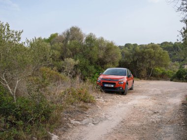Cala Romantica, İspanya - 6 Temmuz 2024: Engebeli bir toprak yolda park etmiş kırmızı bir Citroen arabası yoğun yeşil çalılar ve ağaçlarla çevrili bulutlu bir gökyüzü altında sakin bir kırsal alanda.