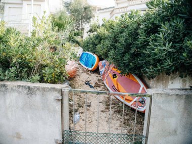 Cala Romantica, İspanya - 6 Haziran 2024: Mallorca 'da yoğun yeşil çalılar ve beton bir duvar arasında dinlenmiş canlı sörf tahtalarının yer aldığı küçük bir kapılı bölge, açık deniz atmosferini çağrıştırıyor.