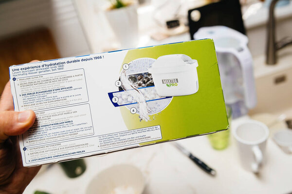 Париж, Франция - 11 августа 2024: Рука, держащая пакет фильтров для воды Maxtra Pro, демонстрирующий информацию о продукте на нескольких языках, с раковиной и кухонными принадлежностями на заднем плане.