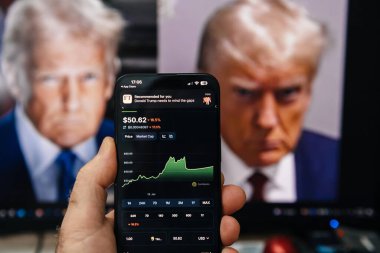 Paris, Fransa - 20 Ocak 2025: Donald Trump 'la ilgili bir tebligat ortaya çıkarken kripto para birimi fiyat tablosunu gösteren bir akıllı telefon. Trump 's portreleri arka plandaki monitörlerde görülebilir.