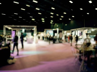 Aydınlatılmış kabinler, dağınık katılımcılar ve canlı ışıklandırma ve etkinliklerle dolu geniş bir kapalı alan gösteren bir ticaret fuarı mekanı bulanık bir görüntü..