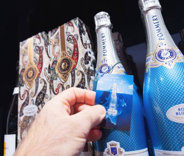 Frankfurt, Germany - Dec 30, 2024: A close-up of a hand holding a tag featuring Pommery Royal Blue Sky champagne, with elegant bottles and ornate packaging on a retail shelf. clipart