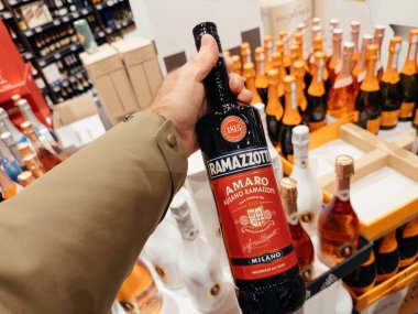 Frankfurt, Germany - Dec 30, 2024: A hand holding a Ramazzotti Amaro Milano bottle in a vibrant liquor store section, surrounded by colorful bottles of champagne and spirits on display. clipart