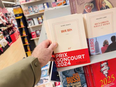 Paris, Fransa - 28 Aralık 2024: Bir el, Kamel Davut 'un Houris adlı kitabını seçerek, iyi organize edilmiş bir kitapçıda, diğer başlıklar da dahil olmak üzere uzun metrajlı edebiyat sergilerini sergiledi..