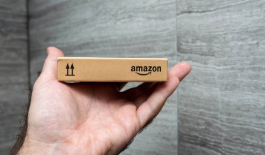 Paris, Fransa - 22 Kasım 2021: Küçük bir Amazon markalı karton kutuyu tutan bir elin yukarı doğru ok sembolüyle desenli gri duvar arkaplanına yakın çekimi, ambalaj detaylarını gösteriyor.