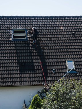 Frankfurt, Almanya - 15 Haziran 2023: Koruyucu giysiler giyen ve kırmızı güvenlik halatı ile korunan bir işçi, arka planda açık mavi gökyüzü bulunan, tavan penceresi yakınındaki fayanslı bir çatı üzerinde bakım yapıyor..