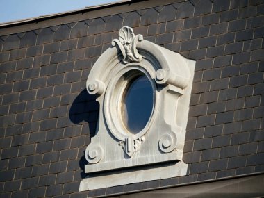 Karmaşık heykel detaylarıyla dekoratif oval yatakhane penceresi, güneş ışığını yansıtan ve klasik mimari cepheye gölge düşüren karanlık bir tahta çatıya kurulur..