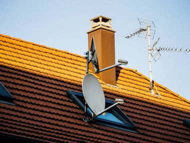 Bir uydu anteni, iki televizyon anteni ve açık mavi gökyüzünün altında bir bacası olan terakotta döşeli bir çatı. Güneş ışığı gölgeler oluşturuyor, geometrik çatı tasarımını vurguluyor.