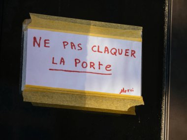 Siyah kapıya bantlanmış el yazısıyla yazılmış, Fransızca kapıyı çarpmamayı öğreten bir tabela. Kırmızı mürekkeple yazılmış mesajın altı çizilmiş ve nezaketen Merci 'yi de içeriyor..