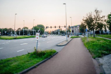 Haarlem. Hollanda - 26 Ağustos 2019: Günbatımında Haarlem 'de geniş bir kavşak, kırmızı parlayan trafik ışıkları, hareket eden arabalar ve yumuşak bir gökyüzü altında çimenli bir yol kenarında çalışan bir bisiklet şeridi -