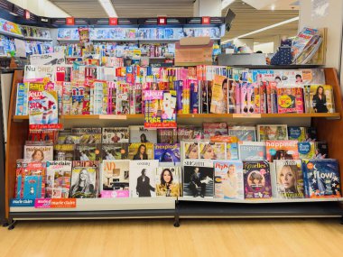 Paris, Fransa - 12 Aralık 2025: Moda ve yaşam tarzı da dahil olmak üzere çok çeşitli başlıklarla iyi ışıklandırılmış bir kitapçıda veya gazete bayiinde katmanlı raflarda sergilenen Fransız ve uluslararası dergilerin renkli gösterimi