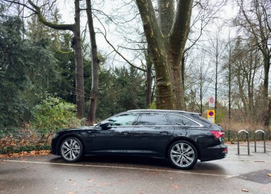 Paris, Fransa - 16 Ocak 2025: Siyah bir Audi steyşın vagonu, sessiz bir park alanındaki uzun yapraksız ağaçların yanında ıslak bir yola park edilmiş ve nemli sonbahar yaprakları huzurlu çevrenin etrafına saçılmıştır.