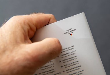Paris, Fransa - 10 Mart 2025: Almanya 'da Yapılan Kalite etiketli ürün ambalajını elinde tutan kişinin yakın plan fotoğrafı, ülke bayrakları ve alan adları bulunan uluslararası AUER Paketleme web siteleri listesinin üzerinde