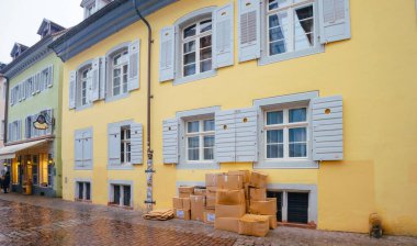 Freiburg, Almanya - 10 Şubat 2025: Düz ve istiflenmiş ayakkabı karton kutuları, atık toplama için bir alışveriş alanının yakınındaki kaldırım taşı sokakta kepenkleri olan sarı bir bina duvarına yerleştirildi
