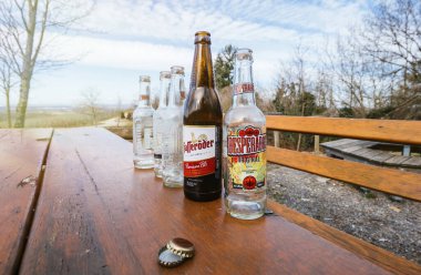 Boş bira şişeleri bir orman ortamında aşınmış ahşap bir masanın üzerinde dizilir, manzaranın manzarası arka plandadır..