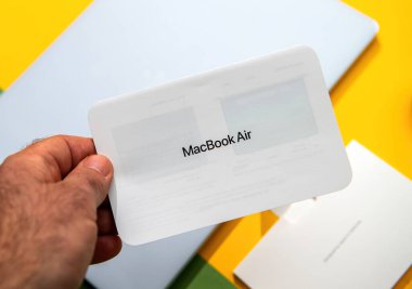 Paris, Fransa - 13 Mart 2025: Bir el, parlak bir çalışma alanının üzerinde görünür cihaz ambalajı, belge klasörü ve canlı sarı ve yeşil yüzey tonlarıyla basılı bir MacBook Air komut kartı tutuyor