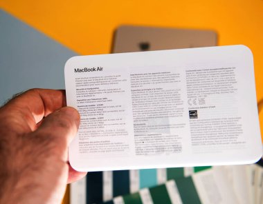 Paris, Fransa - 13 Mart 2025: Elde tutulan bilgilendirme belgesi renk örneklerinin üzerine yerleştirilen MacBook Air için çok dilli güvenlik, uyum ve model özellikleri ve yaratıcı bir çalışma masası içeriyor