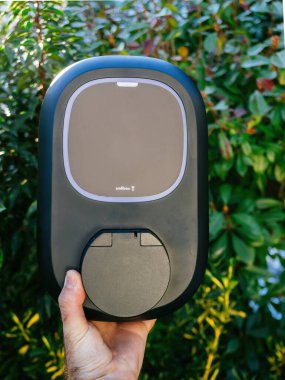 Paris, Fransa - 3 Mart 2025: modern bir Wallbox elektrikli araba şarj aletini yemyeşil bahçe zeminine karşı elinde tutuyor, gösterişli, kullanıcı dostu tasarımı.