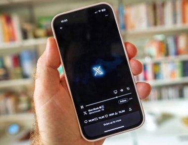 Paris, Fransa - 24 Haziran 2023: Bir erkek, Elon Musk 'ın X platform profilini takip eden istatistiklerle bulanık bir kütüphane geçmişine karşı gösteren bir akıllı telefon tutuyor.