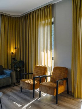 İki kahverengi kadife koltuklu lüks iç mekan altın perdeli bir pencereye bakıyor, zarif ve rahat bir yaşam alanı sergiliyor..