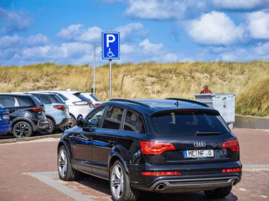 Egmond aan Zee, Hollanda - 25 Ağustos 2024: Aralarında siyah bir Audi SUV 'nin de bulunduğu çok sayıda araba Hollanda' nın kıyı kumu bölgesinde, çimenli kum tepeleri ve arka planda mavi bir gökyüzü bulunan bir engelli park levhasının yakınına park edilmiştir..