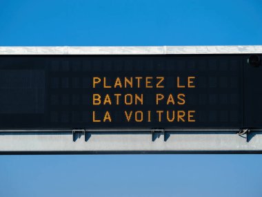 Açık mavi gökyüzündeki dijital bir otoyol tabelası, çevresel bilinci ve alternatif ulaşım yöntemlerini teşvik eden Plantez le baton pas la voiture mesajını göstermektedir.