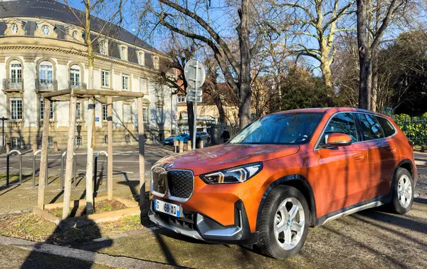 Paris, Fransa - 16 Ocak 2025: Bir Fransız şehir merkezinde güneşli bir caddenin köşesine park edilmiş turuncu bir BMW iX1 elektrikli araç klasik mimari ve kış ağaçlarının yanında uzun gölgeler bırakarak duruyor