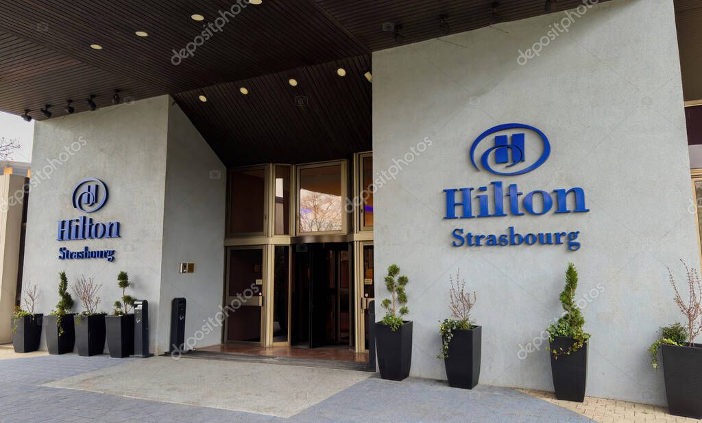 Strasbourg, France - Mar 26, 2025: Main entrance of the Hilton Strasbourg hotel featuring the brand logo on a modern light grey facade with black planters and revolving doors in a luxury setting