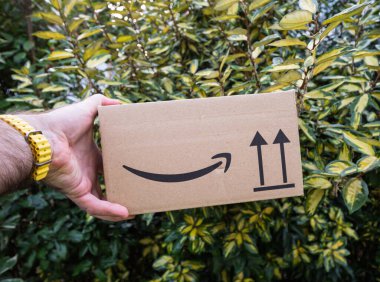 Paris, Fransa - 7 Şubat 2025: Sarı saat takan bir el, açık havada yapraklı bir çalılığın önünde üzerinde Amazon gülüşü logosu ve oryantasyon okları bulunan küçük bir karton paketi tutar.