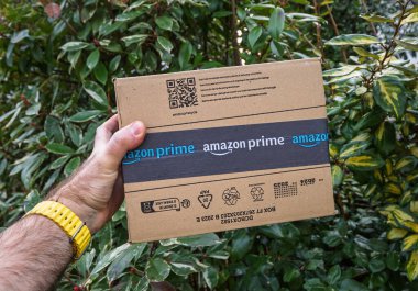 Paris, Fransa - 7 Şubat 2025: Sarı saati olan bir el, yeşil ve sarı bahçe yapraklarının önünde damgalı bant ve geri dönüşüm simgeleriyle mühürlü Amazon Prime karton kutusunu tutar
