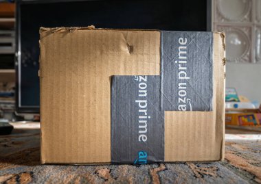 Paris, Fransa - 7 Şubat 2025: Amazon Prime karton teslimat kutusu oyuncak ve elektronik eşyaların yakınındaki bir evin içindeki desenli bir halının üzerinde duruyor.