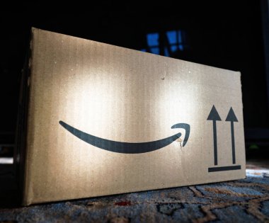 Paris, Fransa - 7 Şubat 2025: Önde gelen Amazon logosunun bulunduğu karton nakliye kutusu ve desenli bir halıya yerleştirilmiş hafif gölgeler ve soluk bir pencere yansıması olan dik oklar