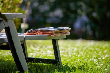 Pembe iş dünyasının finansal ve parlak dergileri güneşli bir bahçedeki şezlongda dinleniyor. Huzurlu bir lüks ve varlıklı boş zamanların rahat temposunu çağrıştırıyor.