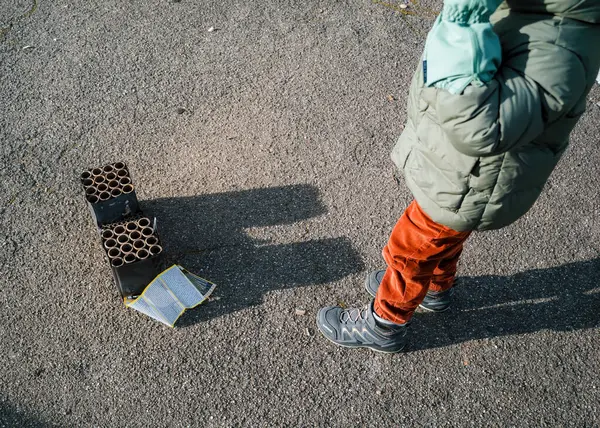 Paris, Fransa - 1 Ocak 2025: Yeşil ceketli ve kırmızı pantolonlu çocuk, asfalt zeminde boş bir havai fişek bataryasının yanında duruyor ve kaldırımda uzun bir gölge oluşturuyor.