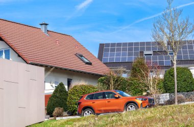 Berlin, Almanya - 2 Mart 2025: Utah Orange BMW iX1 elektrikli SUV, bir Alman köyündeki konut evlerinin önüne park edilmiş, bir çatı katında güneş panelleri ve açık mavi gökyüzü bulunuyor