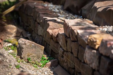 Kırmızı Vosges kumtaşından yapılmış kuru bir duvara yakın çekim, düzensiz şekilli blokları iple dikkatlice hizalanmış, çakıl ve küçük yeşil bitkilerle çevrili,