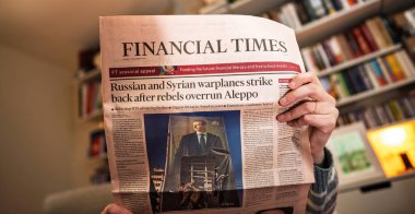 Paris, Fransa - 9 Aralık 2024: Financial Times gazetesinin açık bir kopyasını kitap raflarıyla dolu rahat bir odada tutan bir kişi Halep 'te Rus ve Suriye hava saldırıları hakkında ön sayfada bir makale yayınladı.