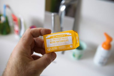Paris, Fransa - 9 Mart 2025: Bir el, arka planda bulanık arkaplanda diş fırçası, fincan ve diğer hijyen ürünleriyle birlikte banyo lavabosunun yanında Dr. Bronner 'ın portakal portakallı doğal sabunlarından oluşan bir çubuğu tutuyor.