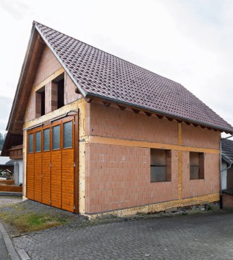Kısmen inşa edilmiş tuğla bir garaj ya da bir Alman köyünde fayanslı çatı ve ahşap garaj kapıları olan, bulutlu bir gökyüzünün altında asfalt bir cadde boyunca kurulmuş bir ek bina.