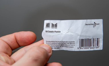 Paris, Fransa - 10 Mart 2025: Bir el kırışık bir DeutschlandCard kuponu tutuyor ve Edeka ve Marktkauf logoları ile tarafsız gri arka planda görülebilen bir barkod sunuyor