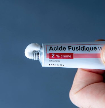 Paris, Fransa - 10 Mart 2025: Bir el, temiz bir mavi arkaplana karşı namludan çıkan% 2 kremli Acide Fusidique metal tüpünü sıkıyor.