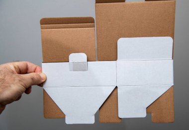 Bir el düz, katlanmış, kahverengi ve beyaz yüzeyli karton bir kutu tutuyor, önceden kesilmiş flapları, katları ve yapıştırılmış bir sekmeyi gösteriyor, paketleme kullanımı için monte edilmeye hazır..