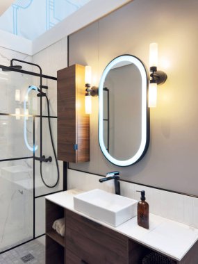 Dikdörtgen damar lavabosu, aydınlatılmış oval ayna, entegre deposu olan ahşap kibir ve siyah çerçeveli cam duş içeren modern banyo görüntüsü.