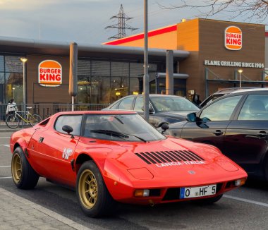 Offenburg, Almanya - 5 Nisan 2025: Parlak kırmızı klasik Lancia Stratos HF modern araçların yanına park etmiş altın tekerlekler Almanya 'da bir Burger King restoranının önünde altın saat ışıklandırması sırasında