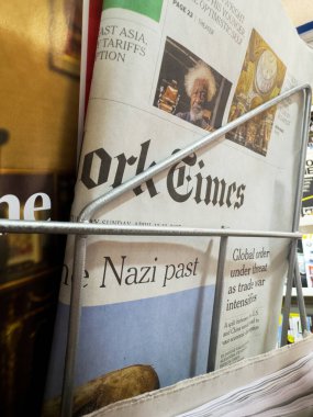Dijon, Fransa - 12 Nisan 2025: Katlanmış New York Times gazetesi, renkli uzun metrajlı resimlerin yanı sıra Nazi tarihi ve küresel düzen tehditleri hakkındaki başlıkları gösteren bir büfede tel askıda oturuyor